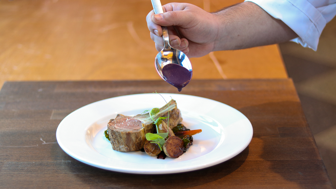 A hand plating a lamb chop with a blueberry sauce.
