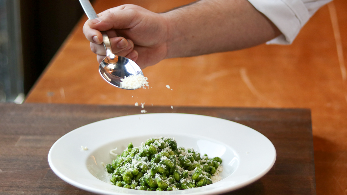 A hand sprinkling cheese over  a dish of Cavatappi Pasta with a Bloomsdale Spinach and Hazelnut Pesto.