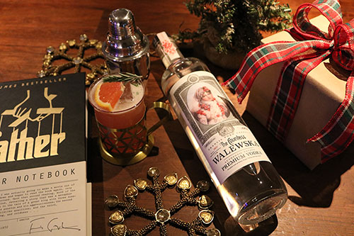 A bottle of Countess Walewska vodka laying down next to a wrapped gift box, a book about the Godfather film, a cocktail shaker and a garnished glass of the Blood Orange Sparkler cocktail.