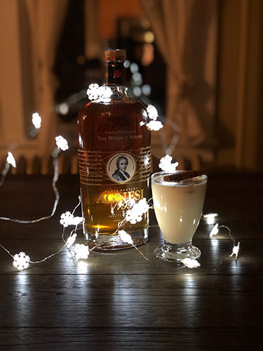 A cream colored Tom & Jerry cocktail in a glass garnished with a cinnamon stick next to a bottle of Agnesi brandy with a string of lit snowflake shaped Christmas lights.