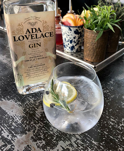 A bottle of Ada Lovelace Gin next to a glass filled with a Gin & Tonic cocktail that is garnished with a lemon slice and rosemary sprig.