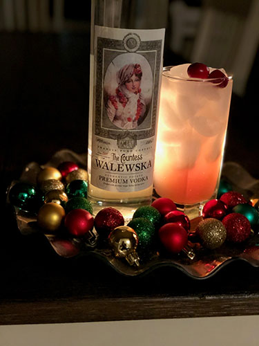 Countess Walewska vodka bottle next to a cocktail in glass that is garnished with cranberries, surrounded by Christmas bulbs.