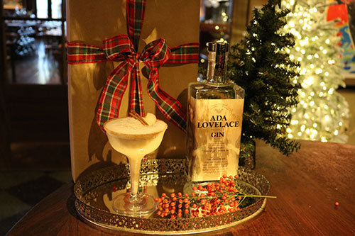 A festive glass of Apple Cider Gin Fizz next to a bottle of Ada Lovelace Gin and wrapped gift boxes.