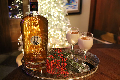 Two glasses of Coquito and a bottle of Agnesi brandy on a tray with an illuminated Christmas tree in the background.