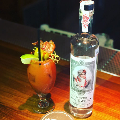 A heavily garnished Bloody Mary cocktail with a lime wedge, olives, and bacon, sitting next to a bottle of Countess Walewska Vodka on a wooden bartop.