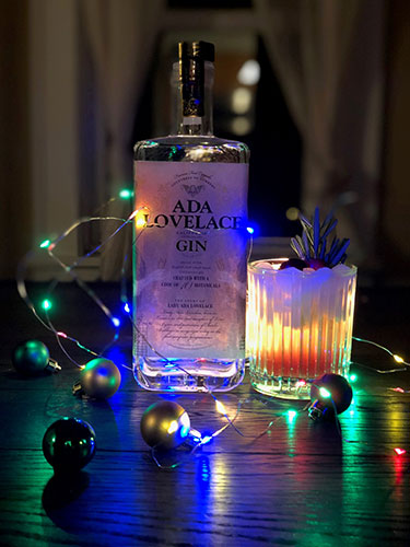 Cranberry Ginger Ale Fizz cocktasil in a glass garnished with a rosemary sprig next to a bottle of Ada Lovelace Gin, surounded by a string of colorful Christmas lights and bulbs.